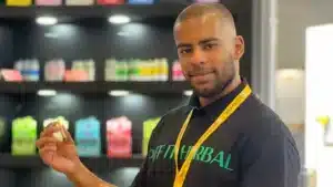 Mann mit schwarzem DIFF HERBAL-Hemd und gelbem Namensband hält kleines Produkt vor Regal.